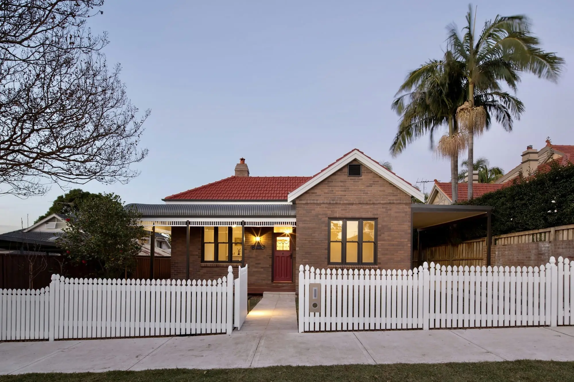 Federation-style brick house with a white picket fence and warm lights at dusk
