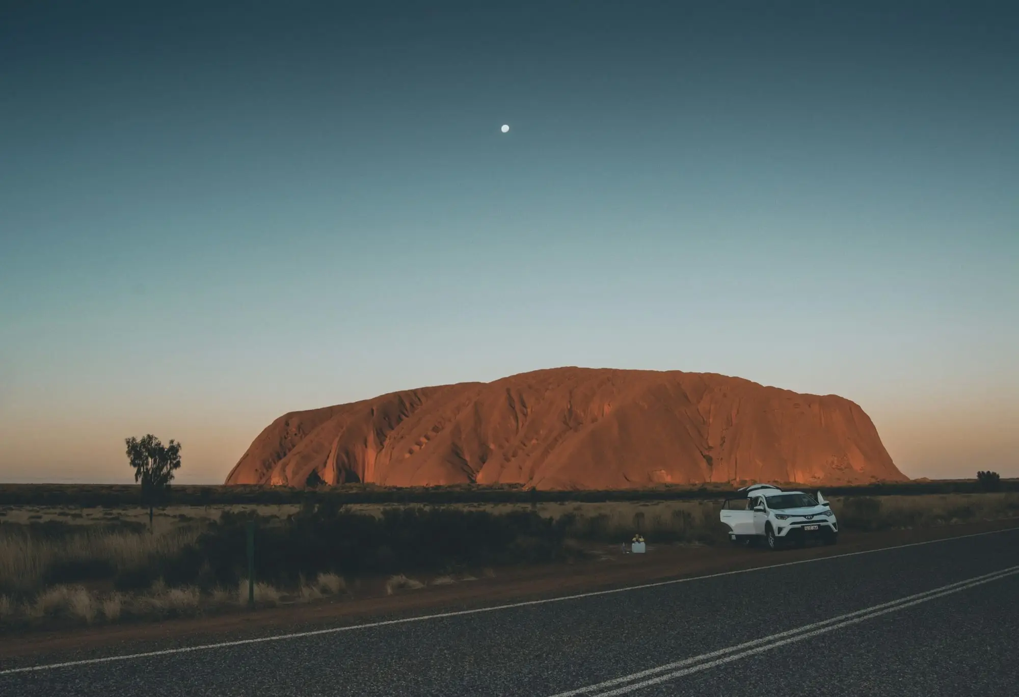 Uluru glowing at dusk with a white car pulled over beside the outback highway